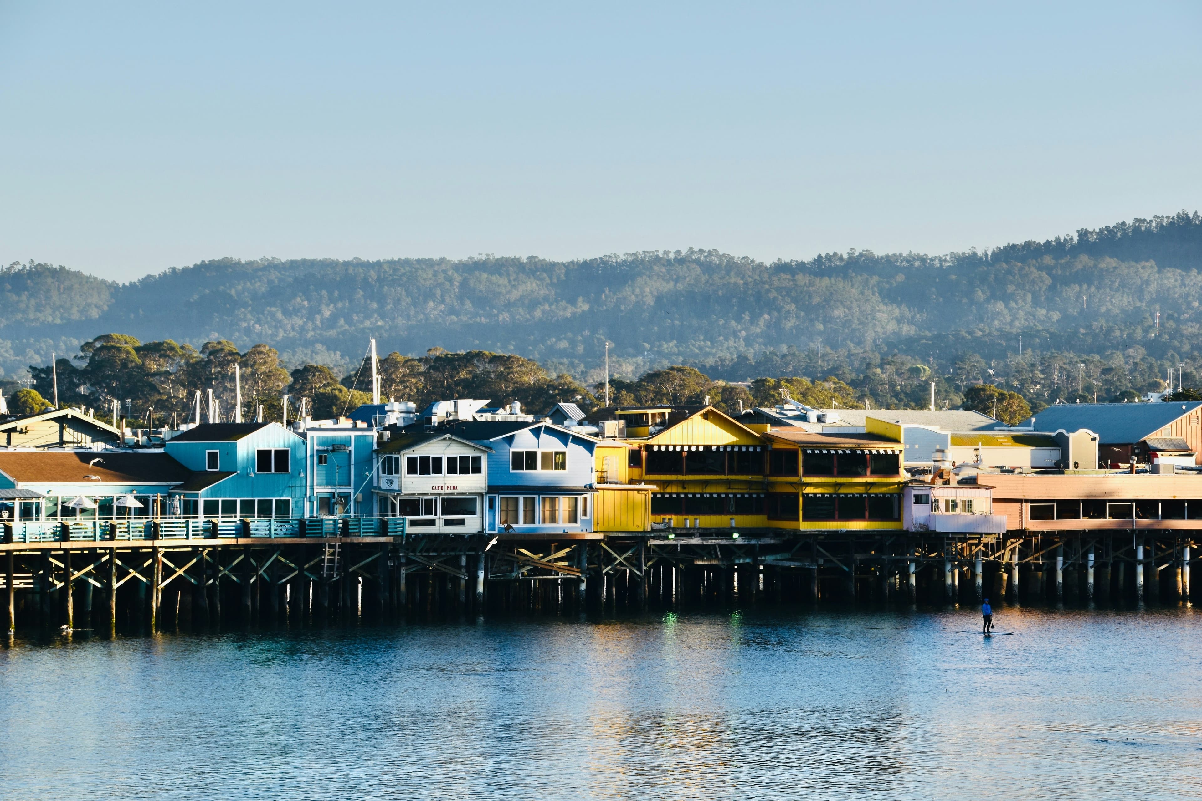 Colorful waterfront buildings line a wooden pier with mountains in the background.