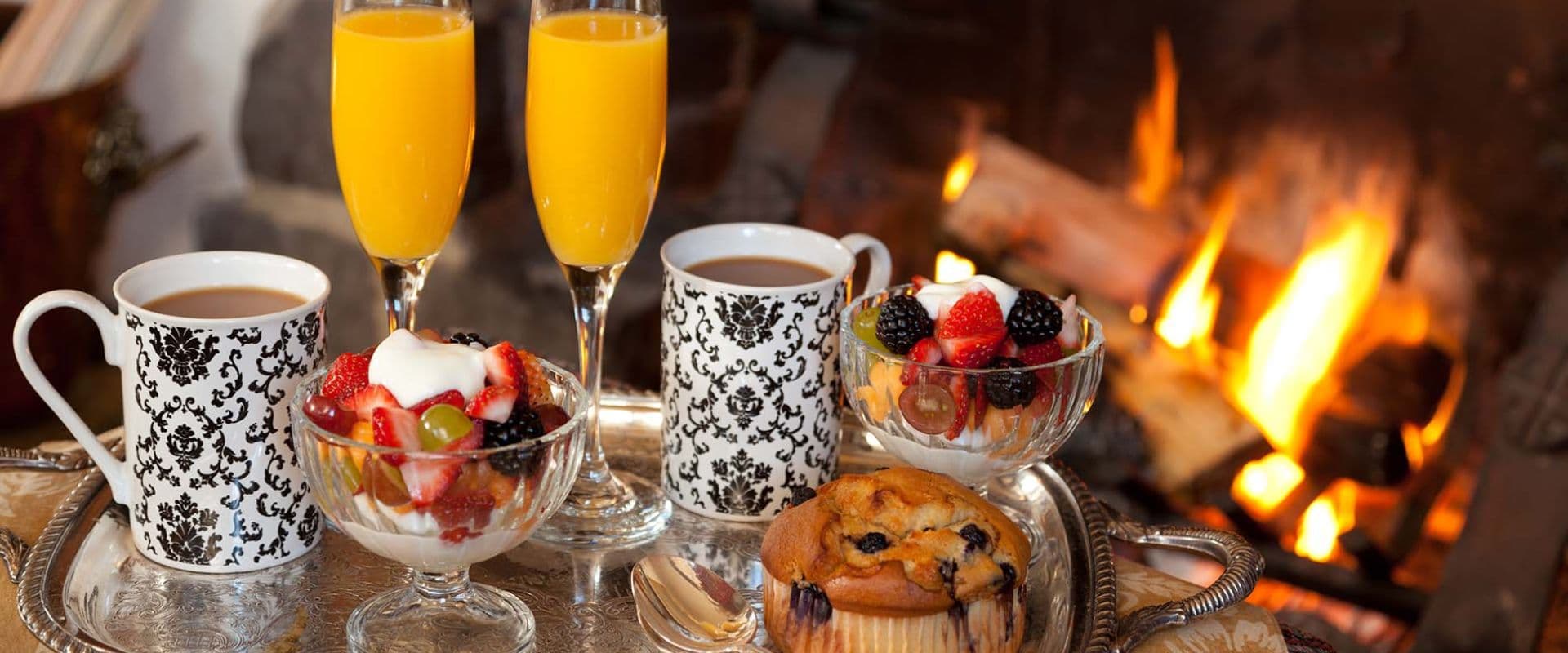 A cozy breakfast spread featuring coffee, orange juice, fruit parfait, and a muffin by a fireplace.