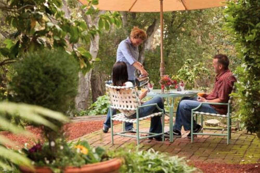 A woman serves coffee to two people seated at a small table under an umbrella in a garden.