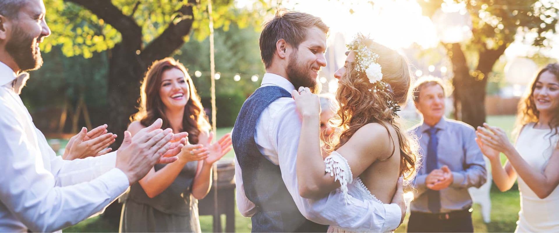 A couple dances joyfully at their wedding while guests applaud in a sunlit outdoor setting.