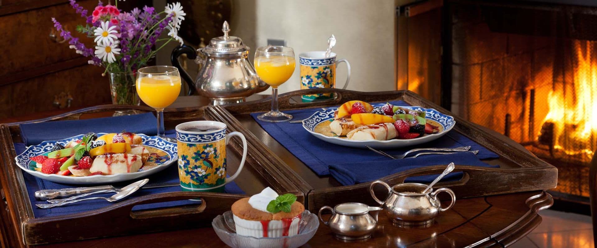 A beautifully arranged breakfast tray with fruit plates, orange juice, and dessert, set near a cozy fireplace.