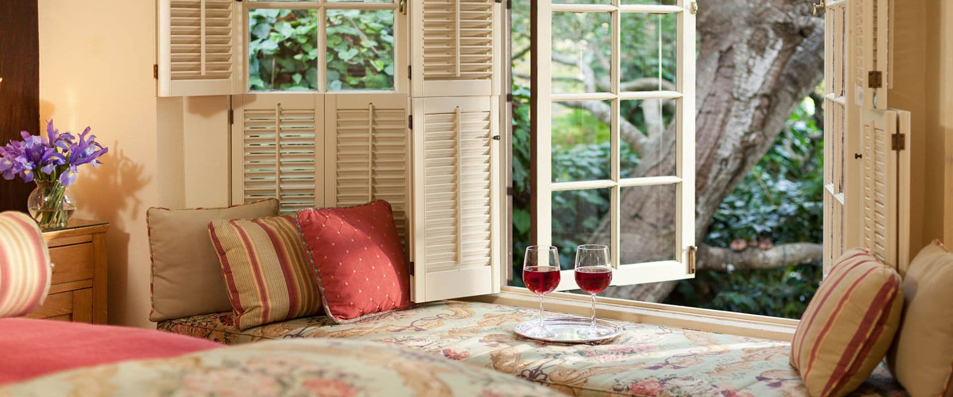 Cozy window seat with decorative pillows and two glasses of red wine, overlooking a lush garden.