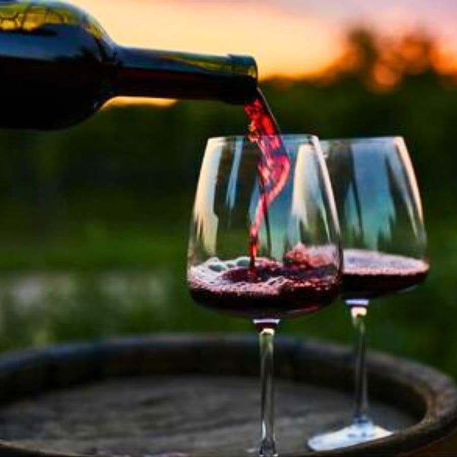 A bottle of red wine pours into two glasses against a sunset backdrop.