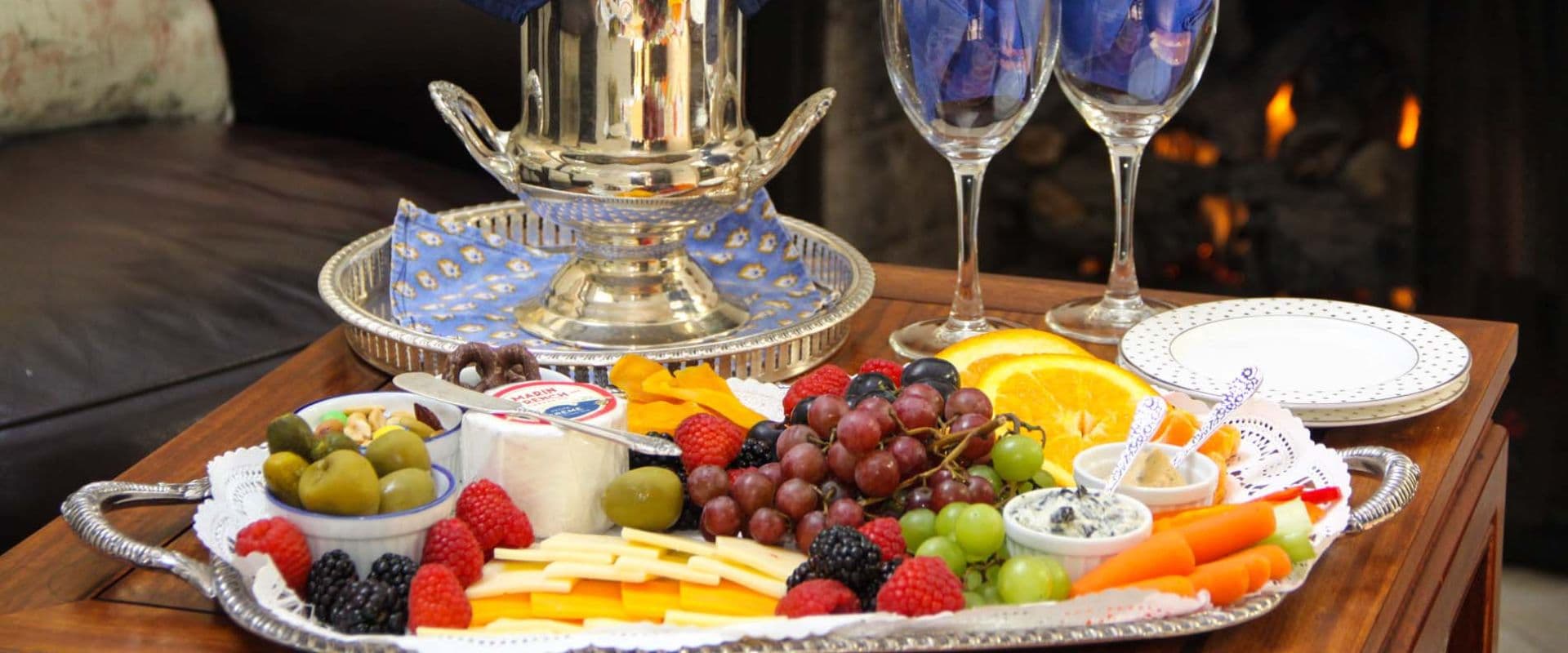 A platter of assorted fruits, cheeses, and olives beside two elegant glasses and a decorative centerpiece.