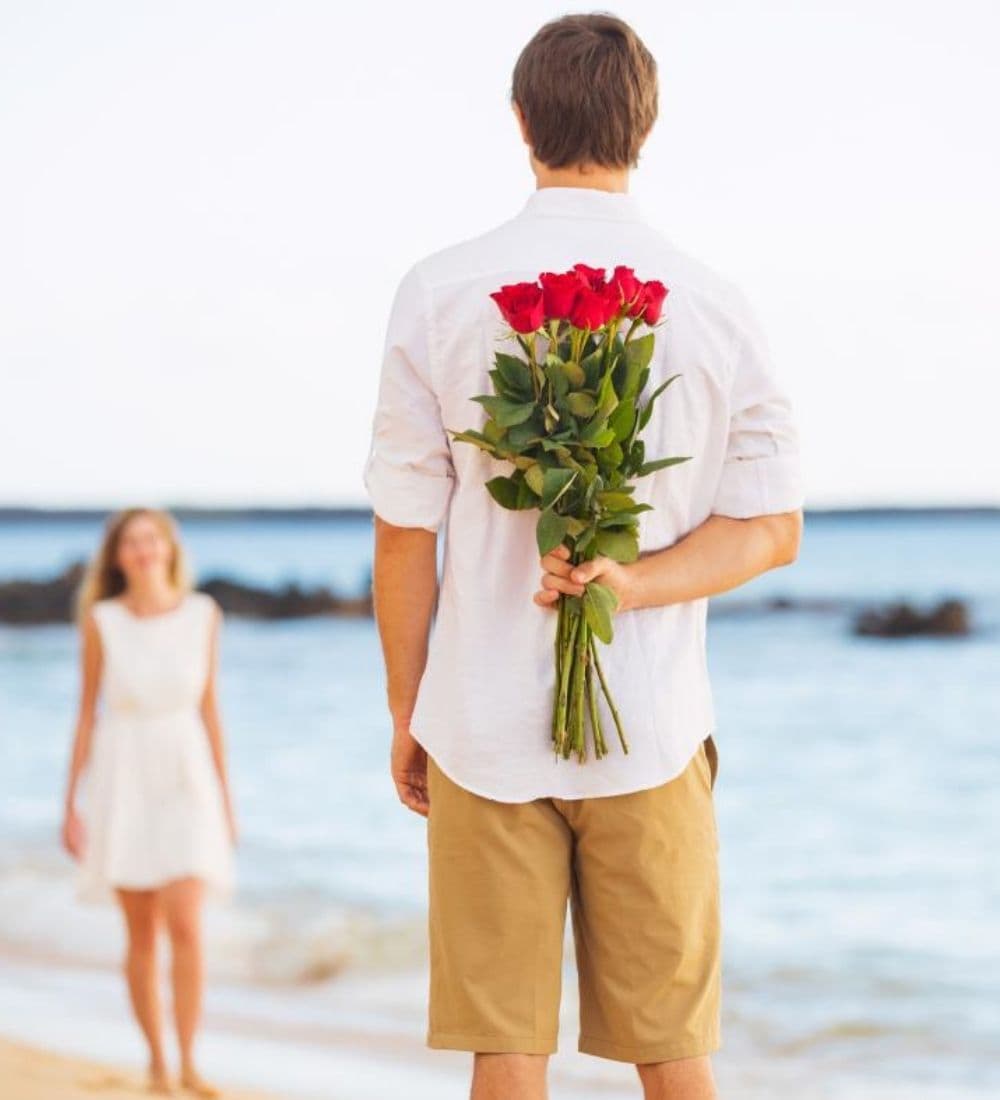 A man stands on the beach holding a bouquet of red roses behind his back while smiling at a woman walking toward him.