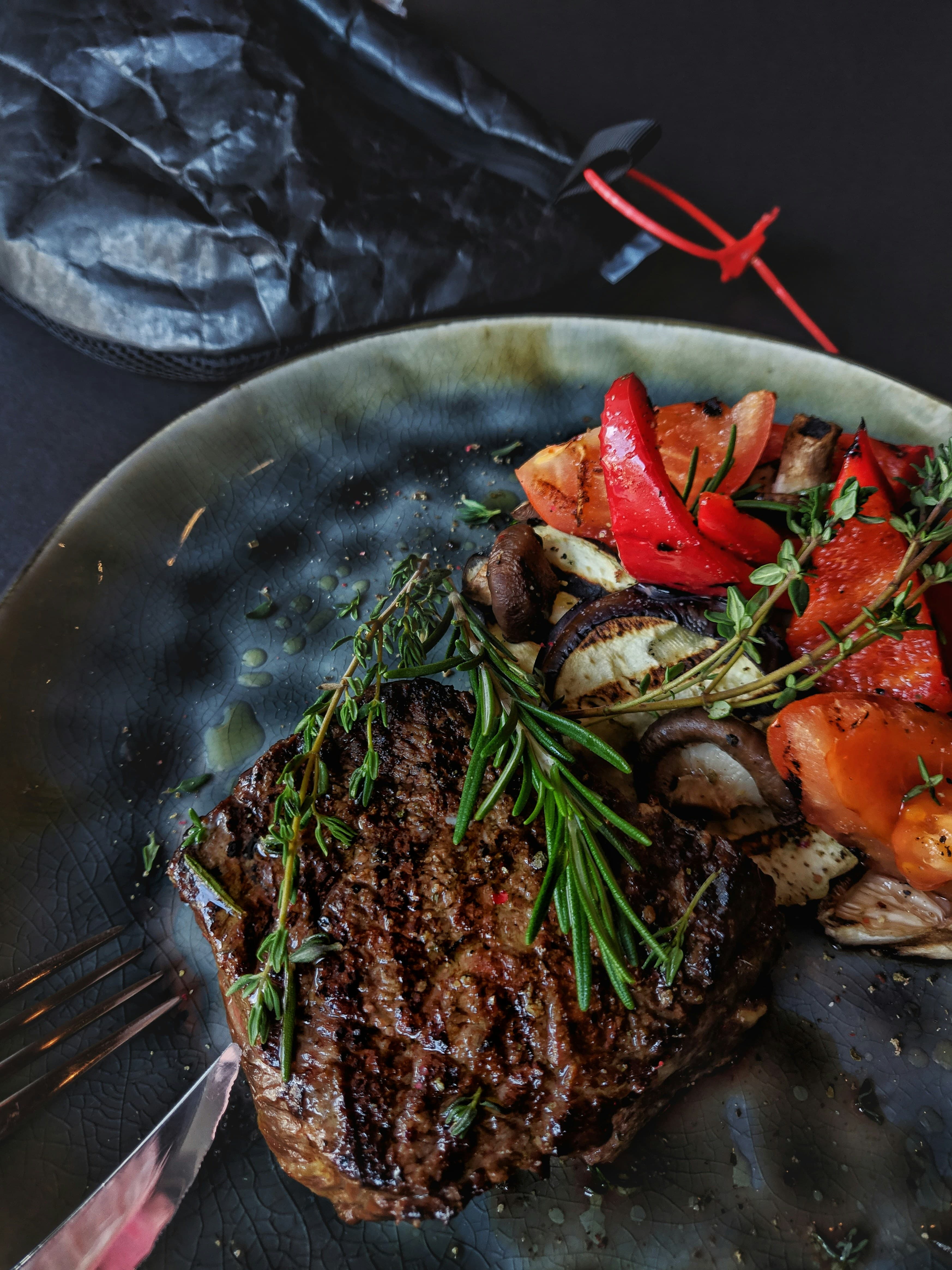 A cooked steak garnished with herbs, accompanied by colorful roasted vegetables on a dark plate.
