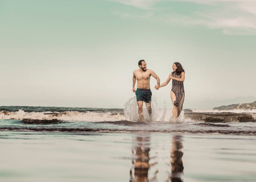 Couple holding hands and running through shallow ocean water with waves splashing around them under a clear sky Couple holding hands and running through shallow ocean water with waves splashing around them under a clear sky