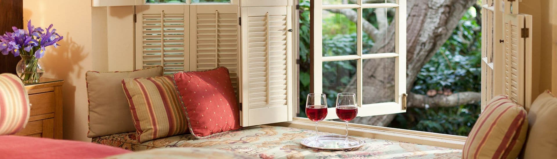 A cozy nook with pillows, two glasses of red wine, and an open window revealing greenery outside.