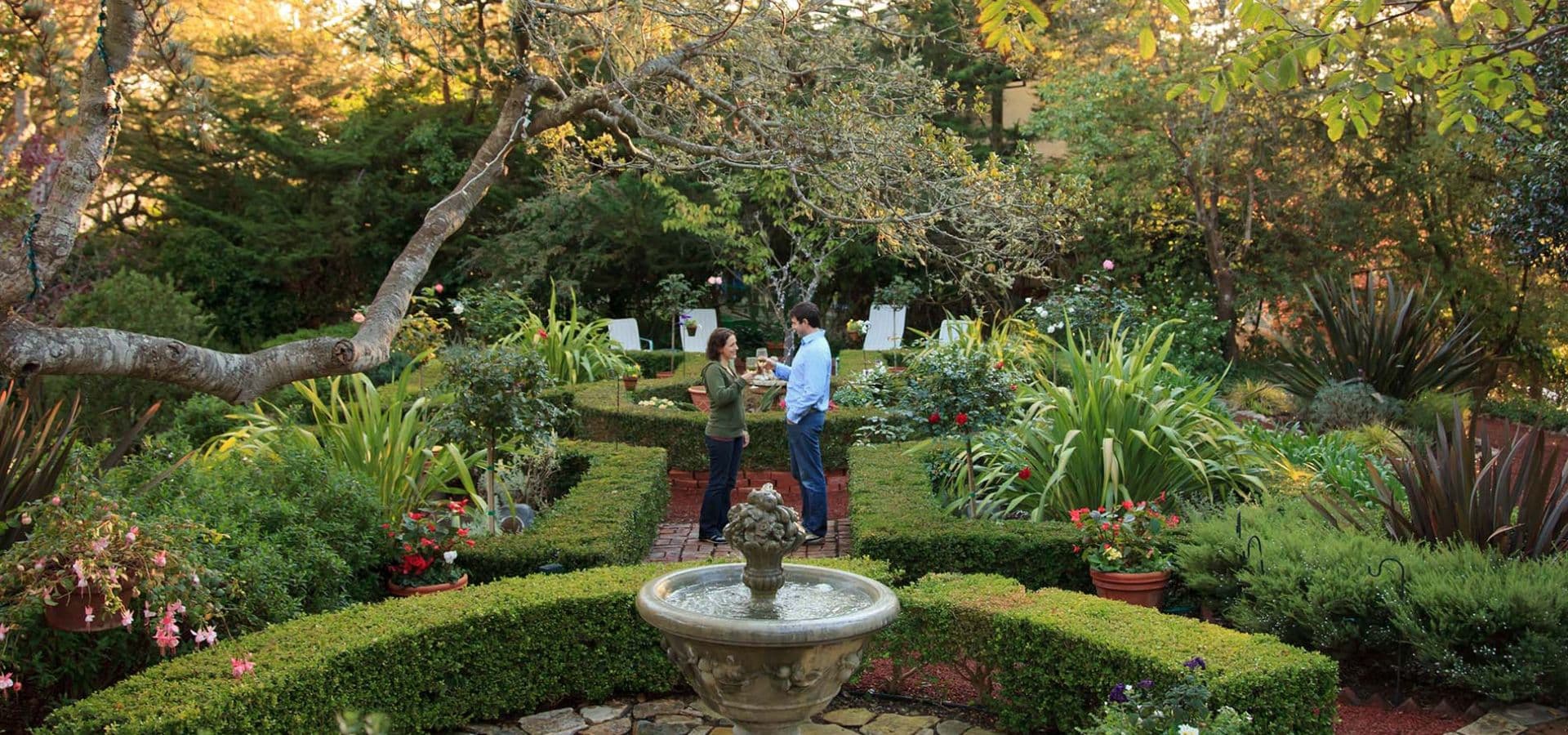 A couple stands in a lush garden featuring a central fountain and vibrant plant life.