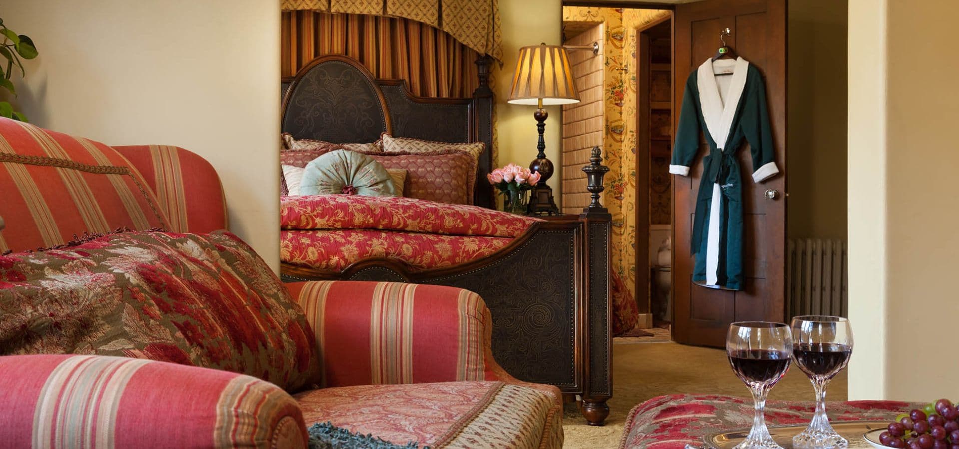 A cozy bedroom with a floral-themed comforter, a lamp, a plush armchair, and two glasses of wine on a table.