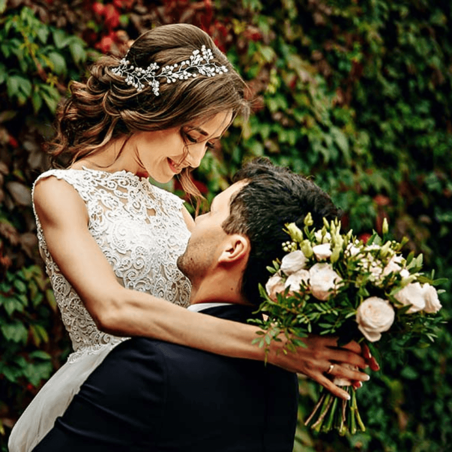 A bride in a lace dress smiles down at her groom while holding a bouquet of flowers.