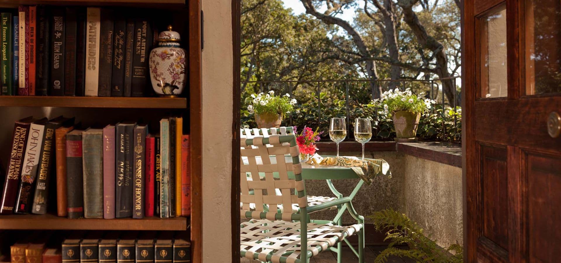 A cozy reading nook with a bookshelf and a doorway leading to a sunlit balcony with two glasses and potted plants.